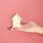 Hand holding a small wooden house model against a soft pink background, symbolizing real estate and property investment.